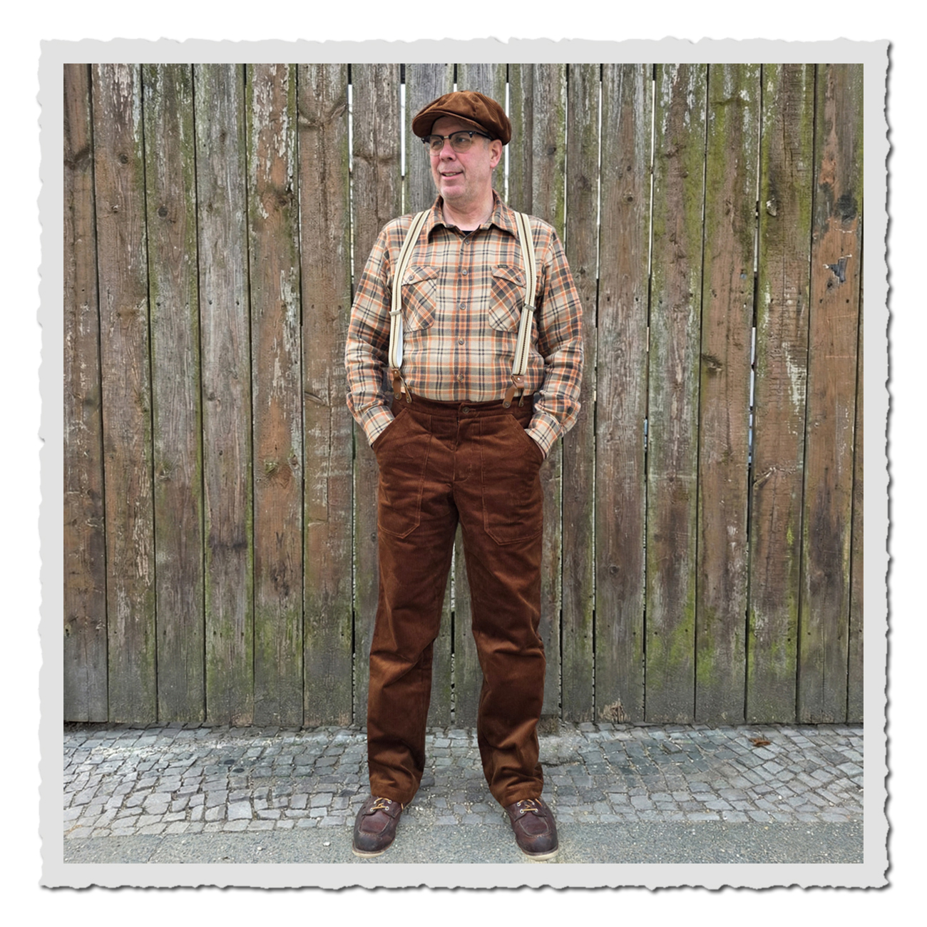 Retronia Arbeiterhose Gustav II in Braun, getragen mit Hosenträgern und Hemd, komplette Vorderansicht einer Person vor Holzzaun.